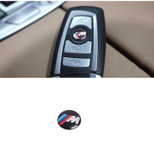 Könnte beinhalten: Nahaufnahme eines schwarzen Autoschlüssels mit einem silbernen Knopf und einem blau-rot-weißen Logo mit der Aufschrift "M".