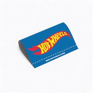 May include: A blue sticker with the Hot Wheels logo in yellow and red. The sticker has a curved shape and the text "WWW.TAGYOURCAR.STORE" at the bottom.