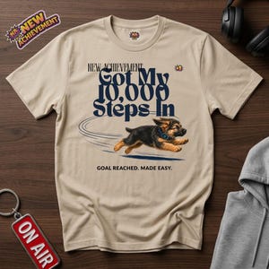 May include: A beige short-sleeve t-shirt featuring a graphic of a running German Shepherd puppy. The text reads "NEW ACHIEVEMENT Got My 10,000 Steps In" and "GOAL REACHED. MADE EASY." The design includes a small "+1" icon and a circular motion trail.