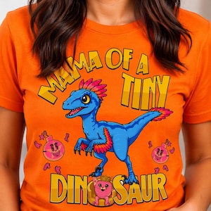 以下が含まれることがあります： 「MAMA OF A TINY DINOSAUR」の文字が入ったオレンジ色のTシャツ。青い恐竜にピンクとオレンジのアクセントが施されたグラフィック。ピンク色の漫画のグラフィックも追加されています。