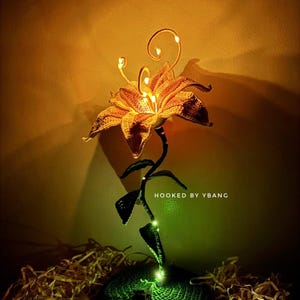 Puede incluir: Una lámpara de flores de ganchillo hecha a mano con luces LED cálidas. La flor es de color amarillo dorado, con detalles en marrón oscuro. El tallo y la base son verdes. El texto "HOOKED BY YBANG" es visible. La lámpara está sobre papel triturado.