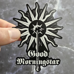 以下が含まれることがあります： 黒と白のステッカー。中央にスパイクのあるオーブと長い柄が付いた、様式化されたサンバーストのデザイン。テキスト「Good Morningstar」は、その下のゴシックフォントで印刷されています。