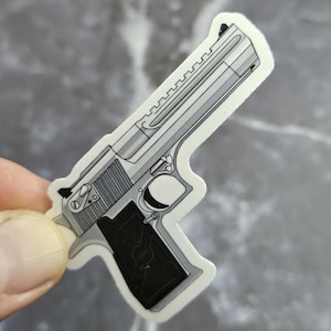 Puede incluir: Una pegatina con una ilustración detallada de una pistola plateada con empuñadura negra. El diseño está delineado en negro, con sombreado para dar un efecto tridimensional. La pegatina se sostiene sobre un fondo gris.