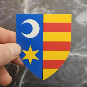 Puede incluir: Un emblema en forma de escudo. El lado izquierdo es azul con una luna creciente blanca y una estrella amarilla. El lado derecho tiene rayas horizontales amarillas y rojas alternas. El emblema está sostenido por una mano.