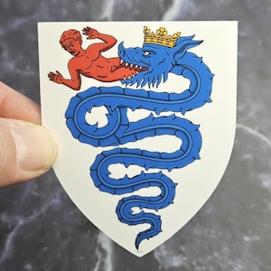 May include: A white sticker featuring a heraldic design. A blue dragon with a gold crown is coiled around a shield. A red figure is positioned above the dragon. The sticker is held by a hand.