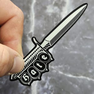 Könnte beinhalten: Ein schwarz-weißer Aufkleber in Form eines Dolches mit dem Wort "Hate" auf dem Griff. Die Dolchklinge ist weiß mit schwarzen Linien. Der Aufkleber wird von einer Hand gehalten.