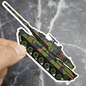 以下が含まれることがあります： 緑、茶、黒の色合いの迷彩柄が施された軍用戦車のダイカットステッカー。戦車には長い砲身、ドイツの国旗の小さなエンブレムが付いた砲塔、そして側面に複数の車輪を備えた目に見えるキャタピラが付いています。