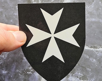 May include: A black shield-shaped sticker featuring a white Maltese cross design. The cross has four arrow-shaped arms, each pointing outwards. The sticker is held by a hand.