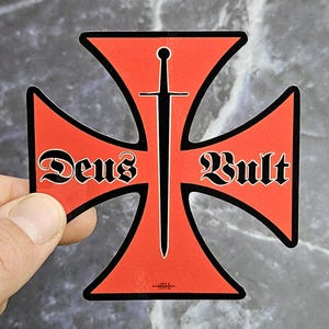 Pode incluir: Um adesivo vermelho em forma de cruz com uma borda preta e um gráfico de espada preta centralizado verticalmente. O texto 'Deus' está impresso em preto no lado esquerdo da cruz, e 'Vult' está impresso em preto no lado direito.
