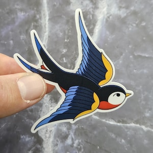 May include: A die-cut sticker featuring a stylized swallow in flight. The bird has a dark blue and black body, bright blue wings with yellow accents, a white face, and a red throat patch. The design is outlined in white with a clean edge.