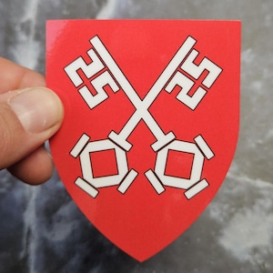 Può includere: Un adesivo a forma di scudo con uno sfondo rosso vivace e un design grafico bianco raffigurante due chiavi incrociate. Le chiavi sono posizionate a forma di X, ognuna con un'impugnatura quadrata e un complesso motivo geometrico all'estremità.