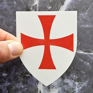 May include: A white shield-shaped sticker featuring a bold red cross pattée centred on the surface. The design displays a classic heraldic emblem with flared arms, presented on a flat, smooth material with clean, sharp edges and a matte finish.