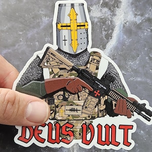 May include: A sticker featuring a cartoon illustration of a knight wearing a helmet with a cross on it, holding a rifle with a red cross on the side. The knight is wearing chainmail and a tactical vest. The sticker has the text "DEUS VULT" in red letters.