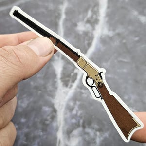 Peut inclure: Un autocollant représentant un fusil avec un canon noir, une crosse en bois marron et un récepteur de couleur or. L'autocollant est tenu entre deux doigts sur un fond gris. Le design du fusil rappelle une arme à levier classique.