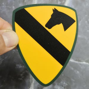 May include: A yellow and black shield-shaped patch with a green border. The patch features a black diagonal stripe and a black horse head silhouette in the upper right corner. The patch is held by a hand.