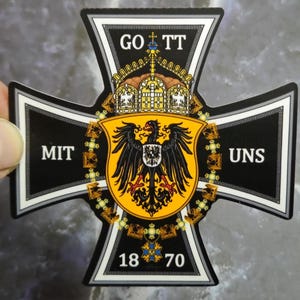 Puede incluir: Una pegatina con forma de cruz en blanco y negro que presenta un escudo central amarillo con un águila negra. El texto "GOTT" aparece en la parte superior, "MIT" a la izquierda y "UNS" a la derecha. Una corona corona el escudo y los números "18" y "70" están en la base.