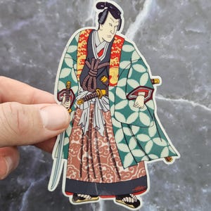 May include: A sticker of a Japanese samurai warrior wearing a green and white patterned robe with a red and white patterned skirt. The samurai is holding two swords and has a serious expression on his face.