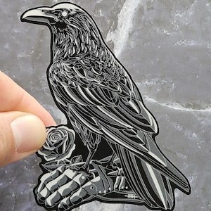 Op de afbeelding: Een zwart-witte sticker met een gedetailleerde illustratie van een raaf die op een metalen handschoen naast een enkele roos zit. Het ontwerp maakt gebruik van contrastrijke lijnen om de veren, de rozenblaadjes en de pantserhandschoen te definiëren.