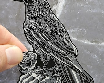 May include: A black and white sticker featuring a detailed illustration of a raven perched on a metal gauntlet next to a single rose. The design uses high-contrast line work to define the bird's feathers, the rose petals, and the armored glove.