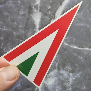 Puede incluir: Una pegatina con un diseño rojo, blanco y verde. La pegatina tiene forma de flecha, con bordes rojos y un interior blanco y verde. La pegatina está sujeta por los dedos de una persona.