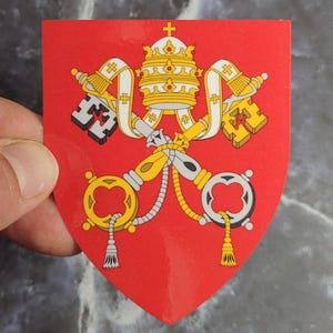 Puede incluir: Una pegatina en forma de escudo con el escudo de armas de la Santa Sede sobre un fondo rojo brillante. El diseño incluye una tiara papal amarilla rematada con una cruz, situada sobre dos llaves cruzadas, una plateada y otra dorada, con cordones blancos.