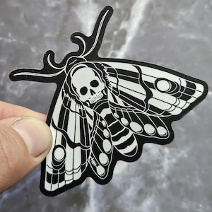 May include: A black and white moth sticker. The moth's head is a skull, and its wings have a detailed pattern. The sticker is held by a hand against a grey background.