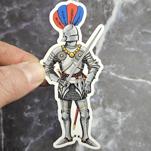 Peut inclure: Un autocollant représentant un chevalier médiéval en armure complète. Le chevalier est représenté avec un casque orné de plumes rouges et bleues, tenant une épée. L'armure est argentée avec des détails dorés.