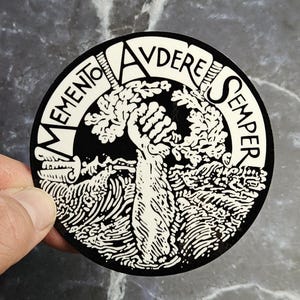 May include: A circular black and white sticker featuring a central graphic of a raised fist emerging from stylised waves. The text MEMENTO AVDERE SEMPER is printed in a bold, curved font along the upper border of the circular design.