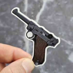 Può includere: Un adesivo fustellato con un'illustrazione dettagliata di una pistola Luger P08. L'arma da fuoco presenta un telaio in metallo nero opaco, una canna lunga e un'impugnatura testurizzata marrone scuro. Il design include dettagli meccanici e un bordo bianco.