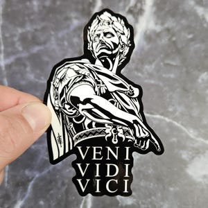 May include: A black and white sticker featuring a stylized illustration of a Roman leader wearing a laurel wreath and armor. The figure is pointing downward toward the text VENI VIDI VICI printed in a bold, white, serif font at the bottom.