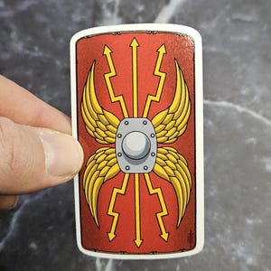 Puede incluir: Una pegatina rectangular con un diseño de escudo romano estilizado. El escudo tiene un fondo rojo con alas amarillas, rayos y flechas. Una placa de metal plateado está en el centro.