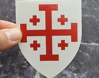 May include: A white shield-shaped sticker featuring a red Jerusalem cross design. The central cross is flanked by four smaller Greek crosses, one in each quadrant. The sticker has a smooth, matte finish and a clean, die-cut edge.