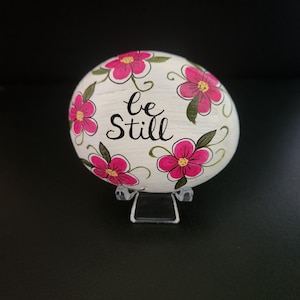 May include: A smooth, oval-shaped white stone painted with vibrant pink flowers featuring yellow centers and green leaves. The words "be Still" are written in black cursive script in the center of the stone, which rests on a clear plastic stand.