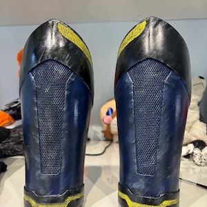 Peut inclure: Deux pièces d'armure de costume bleues et noires avec des détails jaunes. L'armure a une texture en maille au dos et une finition métallique. Le design présente une forme incurvée avec une bande jaune en haut et en bas.