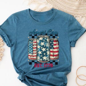May include: Blue t-shirt featuring a graphic of three beverage cans with American flag designs. The text "Ameri Can" is arched above the cans, with "EST. 1776" below. The shirt has rolled-up sleeves.
