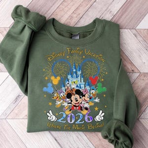 Peut inclure: Un sweat-shirt vert olive avec un graphisme color&eacute; de personnages Disney, un ch&acirc;teau, des feux d'artifice et le texte "Disney Family Vacation 2026 Where the Magic Begins". Le motif comprend Mickey Mouse, Minnie Mouse, Donald Duck et Dingo.