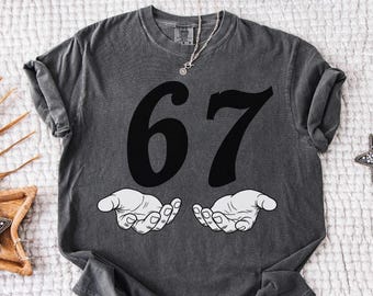 May include: A dark gray t-shirt with the number 67 in large black font. Below the numbers are two white hands in a cupped position. The word "Pepper" is in the top right corner.