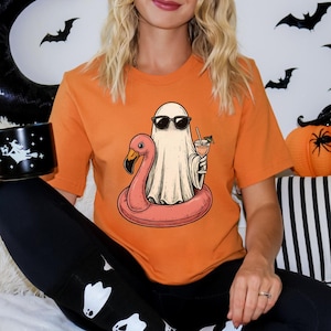 May include: Orange t-shirt featuring a cartoon ghost wearing sunglasses and floating on a pink flamingo floatie, holding a cocktail. The image has a Halloween theme, with bats and a pumpkin in the background. A black mug with a witch design is held in the hand.