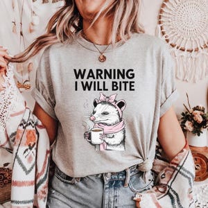 Könnte beinhalten: Ein graues T-Shirt mit einem Aufdruck eines Opossums, das eine Tasse Kaffee hält. Der Text "WARNING I WILL BITE" ist auf dem T-Shirt aufgedruckt.