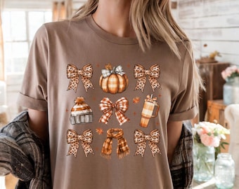 Puede incluir: Camiseta color espresso con un gráfico temático de otoño. El diseño incluye lazos, una calabaza, un gorro, una bufanda y una taza de café. La camiseta es de cuello redondo y manga corta.