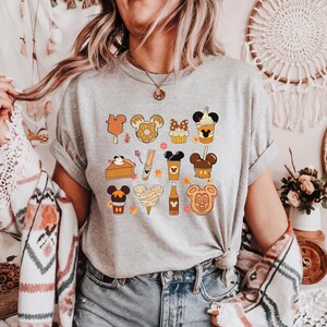 May include: A heather gray t-shirt featuring a collection of cartoon food items with Mickey Mouse ears. The design includes ice cream, donuts, and drinks in shades of brown, orange, and yellow. The shirt is worn with denim shorts.