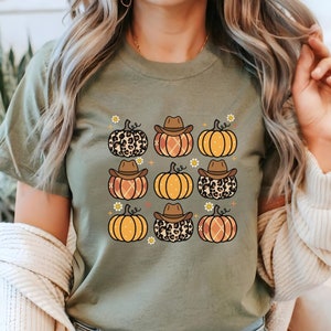 Puede incluir: Una camiseta verde con un patr&oacute;n de calabazas naranjas y marrones que llevan sombreros de vaquero. Algunas calabazas tienen un dise&ntilde;o de estampado de leopardo.