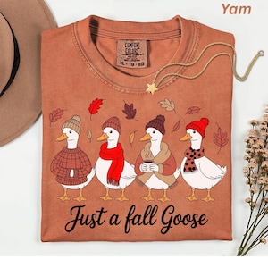 May include: A yam-colored t-shirt featuring a whimsical illustration of four geese dressed in fall attire. The geese are wearing hats, scarves, and sweaters, with the text "Just a fall Goose" below. A gold necklace with a star pendant is draped across the shirt.