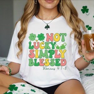 May include: White t-shirt with a green, yellow, pink, and blue graphic that says "Not Lucky Simply Blessed Romans 4:7-8".