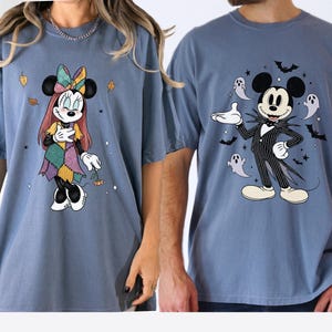May include: Two blue jean-colored t-shirts. One features Minnie Mouse in a patchwork dress with autumn leaves. The other has Mickey Mouse dressed as Jack Skellington, surrounded by bats and ghosts. Both shirts have short sleeves.