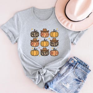 Puede incluir: Una camiseta gris con un patr&oacute;n de calabazas con sombreros de vaquero y estampado de leopardo. Las calabazas son de color naranja y marr&oacute;n con un tallo amarillo.