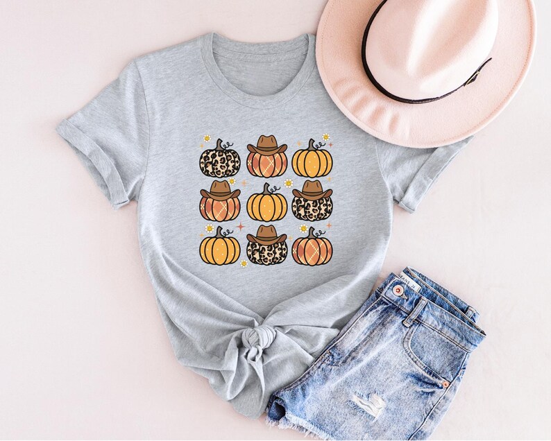 Puede incluir: Una camiseta gris con un patr&oacute;n de calabazas con sombreros de vaquero y estampado de leopardo. Las calabazas son de color naranja y marr&oacute;n con un tallo amarillo.