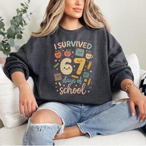 May include: Dark gray sweatshirt with the text "I Survived 67 days of school" in colorful letters and graphics. The design includes smiling apples, books, pencils, and a coffee cup. The sweatshirt is a crew neck style.
