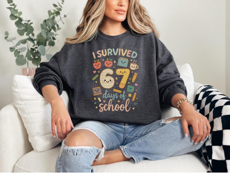 May include: Dark gray sweatshirt with the text "I Survived 67 days of school" in colorful letters and graphics. The design includes smiling apples, books, pencils, and a coffee cup. The sweatshirt is a crew neck style.