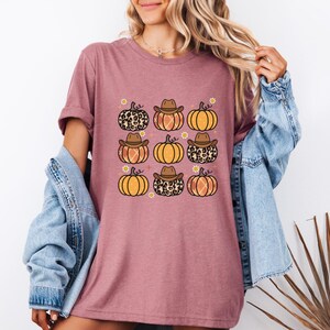 Puede incluir: Una camiseta malva con un estampado gr&aacute;fico de nueve calabazas con sombreros de vaquero. Las calabazas son de color naranja y marr&oacute;n con un estampado de leopardo.